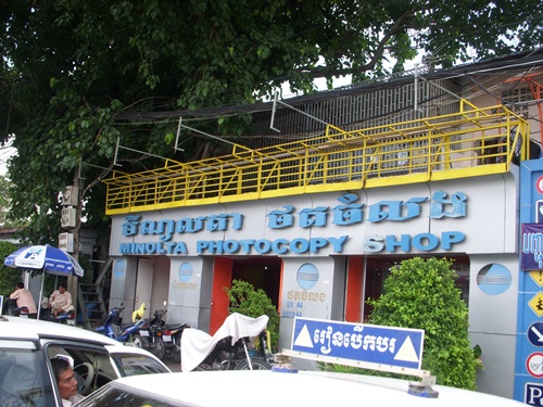 Photo is of a Cambodian printing shop taken from the street.  The shop is a small, white building with blue squiggly Cambodian script that probably says who they are.  Below the squiggly letters is the word also in blue Photocopy Shop.