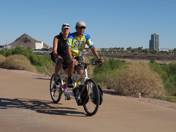 Photo of Tauru and Christi riding on a bike path in Tempe, Arizona with Arizona State University's a mountain in the background. 
