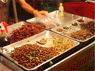 Title: Street Foods - Description: Photo is a street venfor with trays of various foods.  They look like fried insects, but not 100% sure.  Although, this wouldn't be a surprise given how exotic food is in Thailand.  