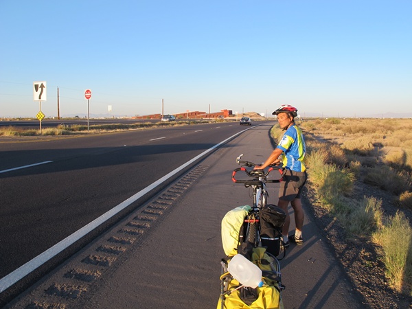 Photo is of Tauru holding the bike while waiting for a break in traffic so we can cross highway 347. It is late afternoon with the sun splashing rays of orange across the desert landscape.